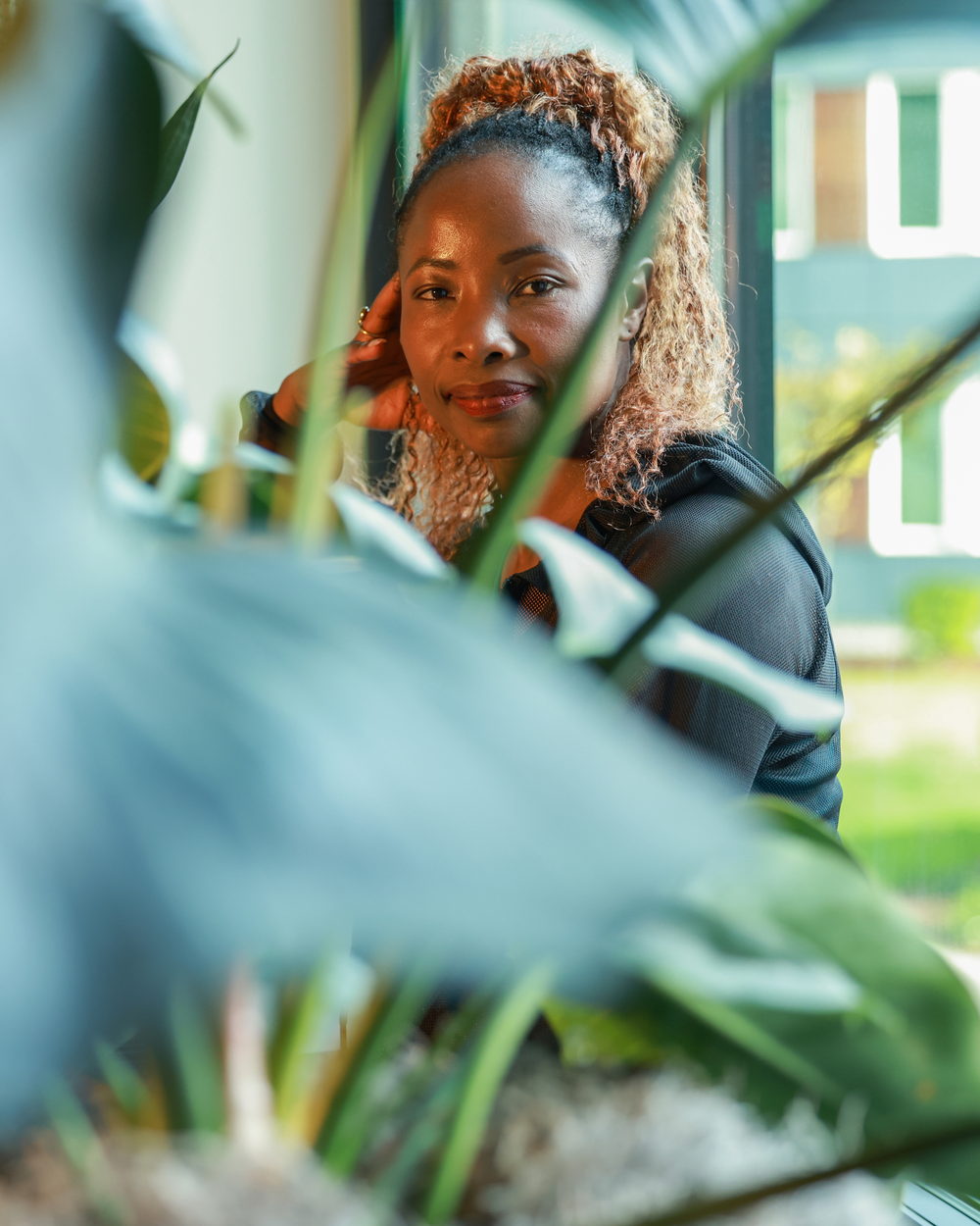 Betty resting her head on her hand, looking at the camera through the leaves of a houseplant