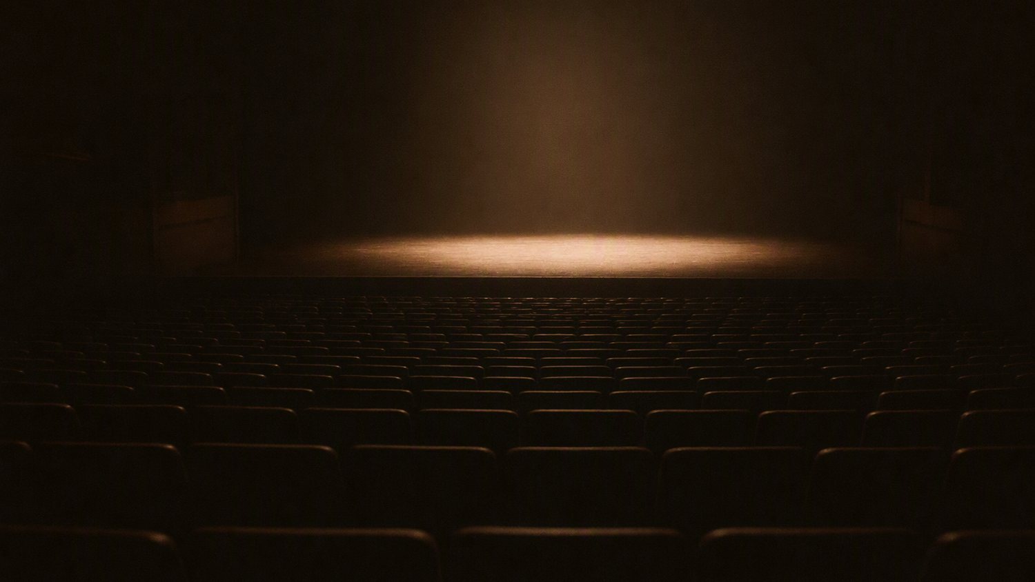 A dark auditorium seen from the back, rows of empty seats, one pool of warm light on a bare stage far away.