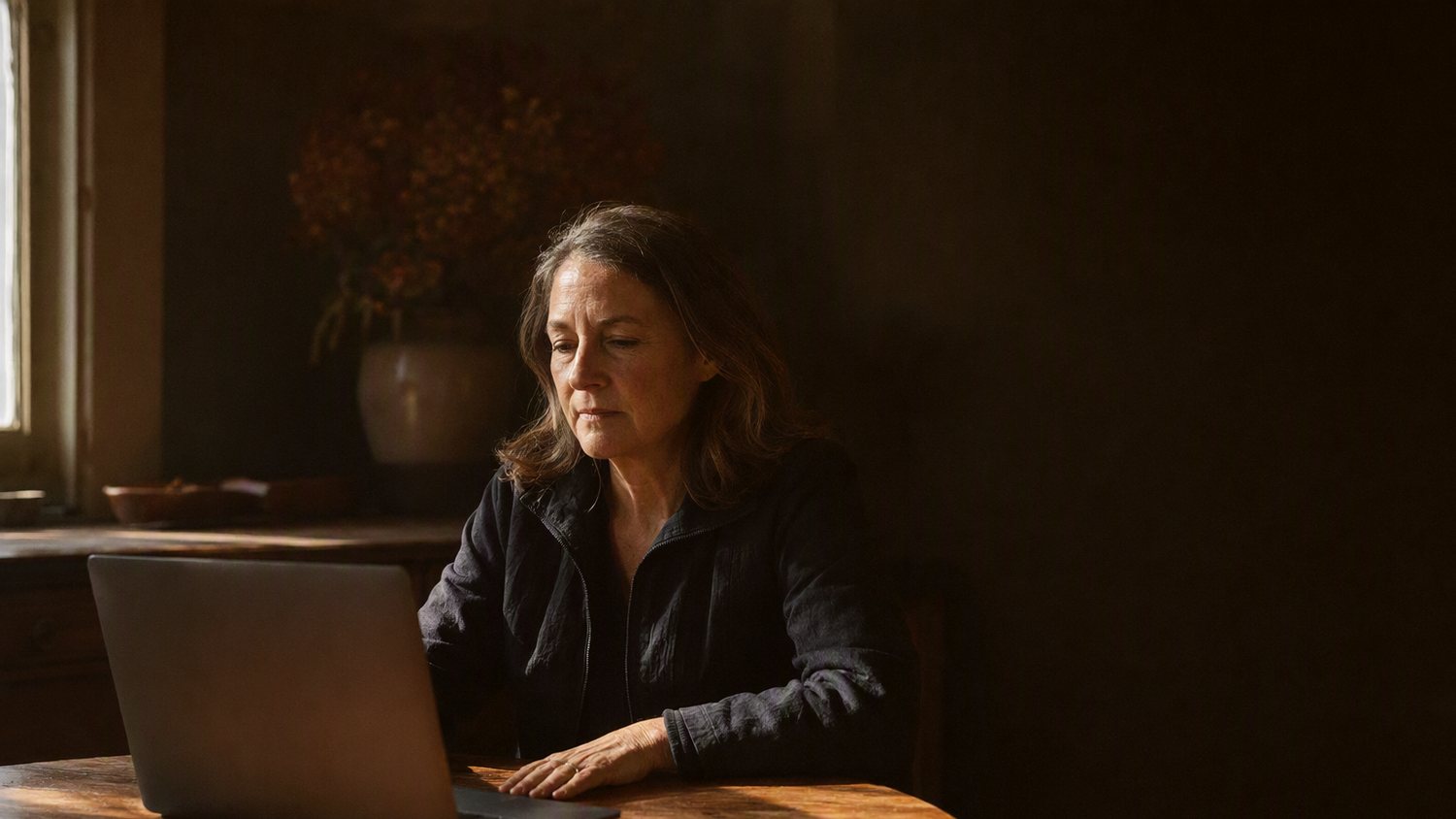 A woman sitting back from a laptop at a warm table, reading something carefully and taking her time.