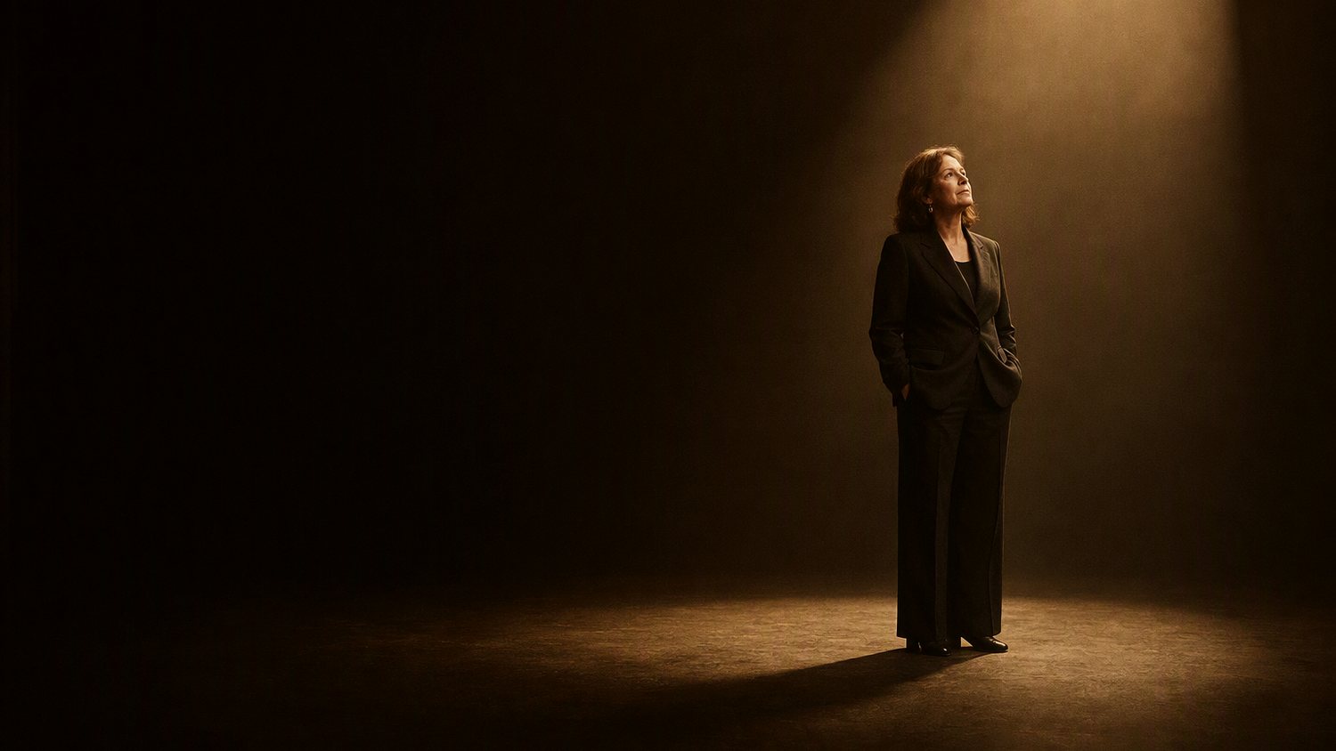 A vast dark room with one shaft of warm light landing as a pool on the floor, and a single figure standing in it.