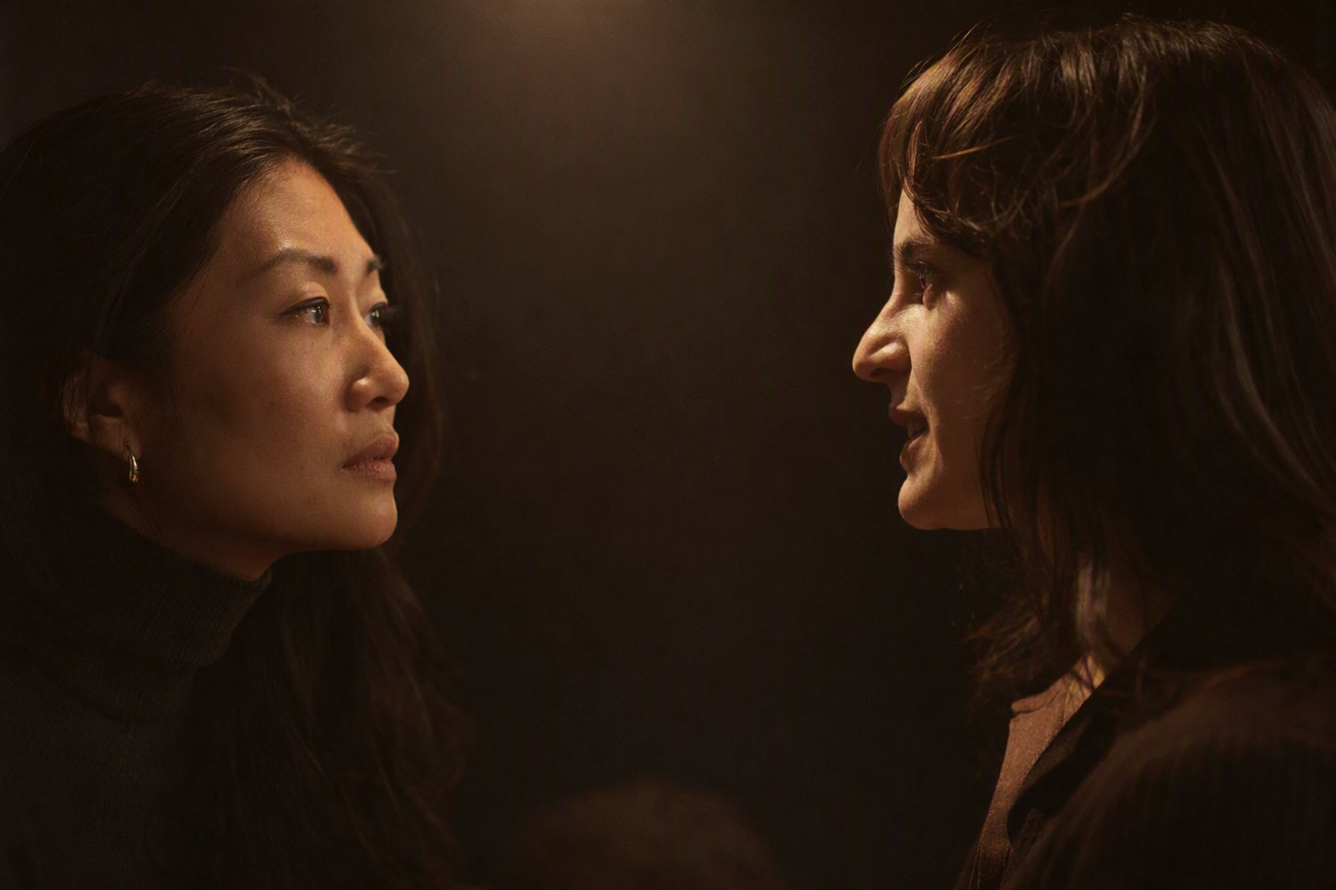 Two women in a dark room mid conversation, one listening closely while the other speaks.