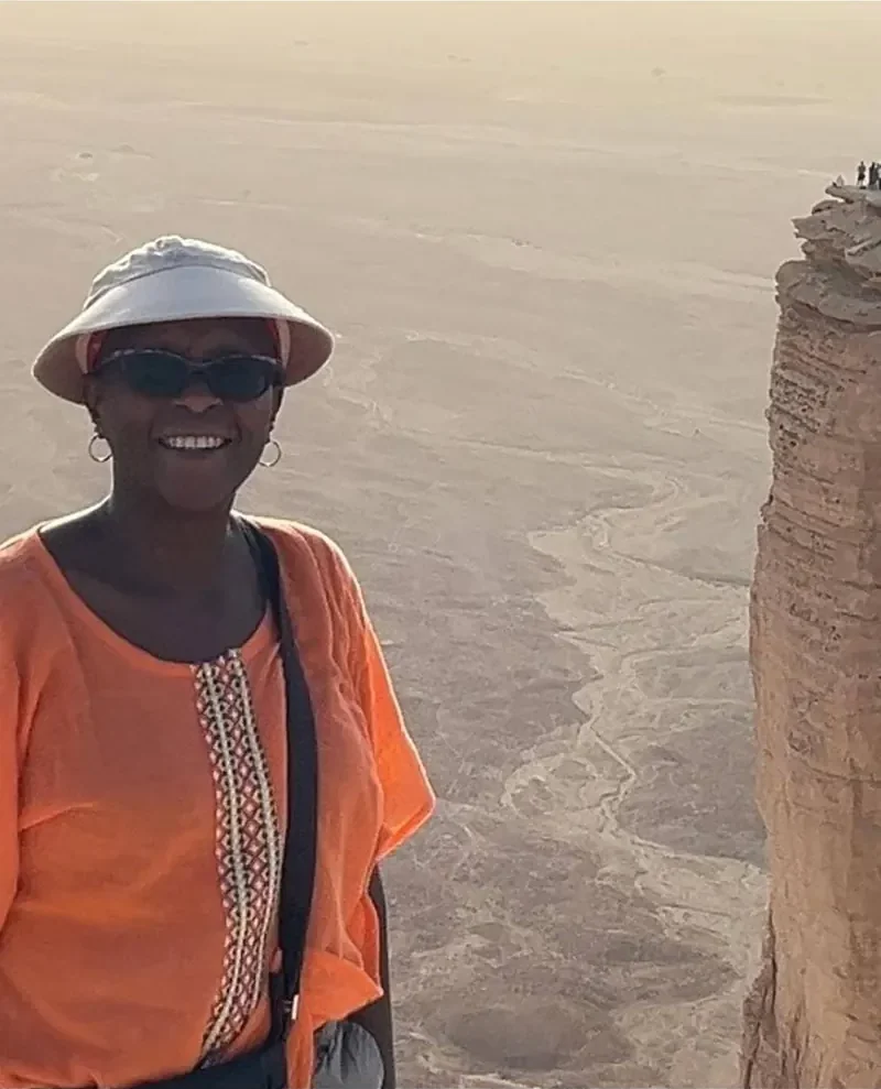 Maria Kamau smiling in an orange top and sun visor at a canyon viewpoint