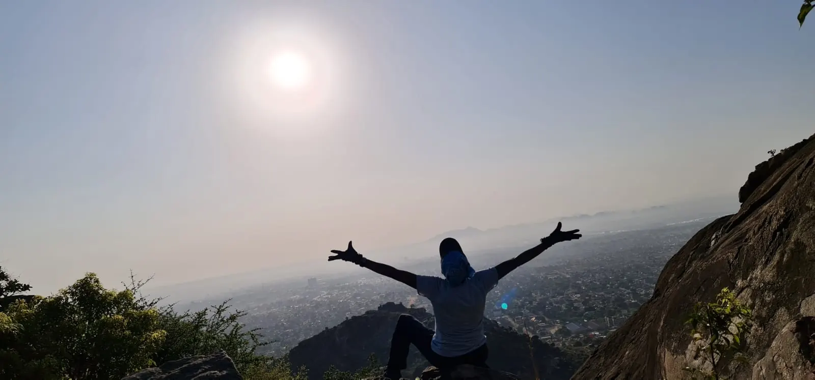 A traveler with arms flung wide on a rocky summit, a whole city spread out below in the morning haze